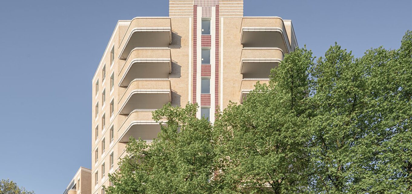 Art Deco style residential brick building with curved balconies and tile details, behind a tree with green leaves