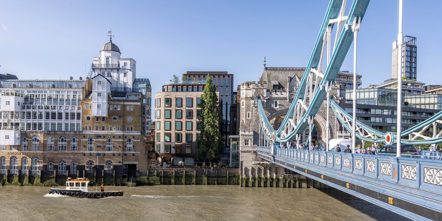 Brick and metal cladded building with reflective windows with the River Thames and Tower Bridge in front.