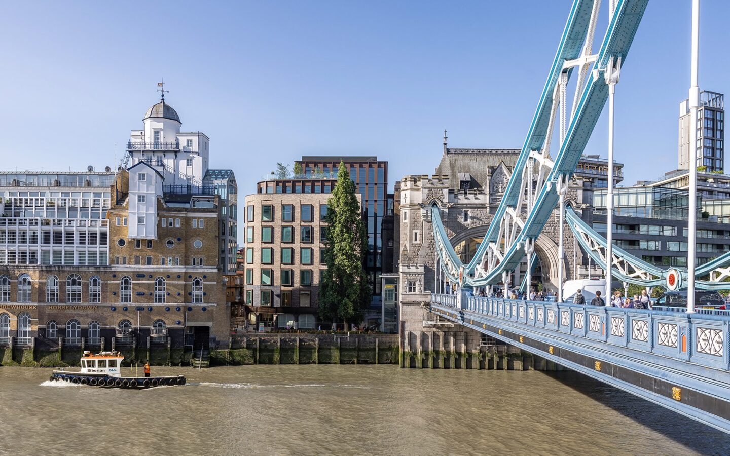 Brick and metal cladded building with reflective windows with the River Thames and Tower Bridge in front.