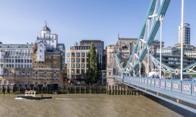 Brick and metal cladded building with reflective windows with the River Thames and Tower Bridge in front.