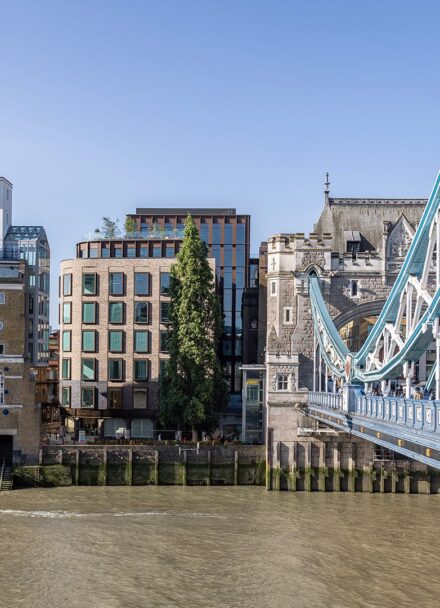 Brick and metal cladded building with reflective windows with the River Thames and Tower Bridge in front.