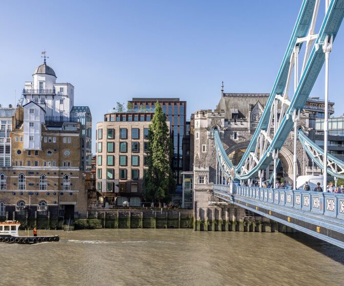 Brick and metal cladded building with reflective windows with the River Thames and Tower Bridge in front.