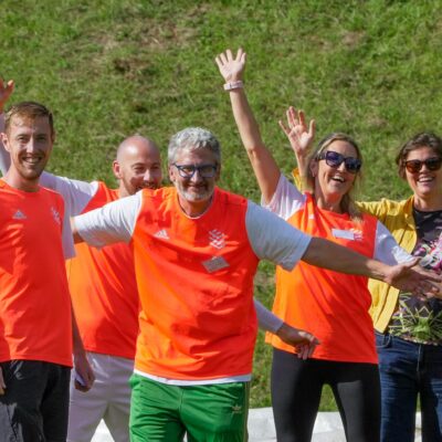 Group of five people waving and gesturing happily, wearing bright orange sports tops.