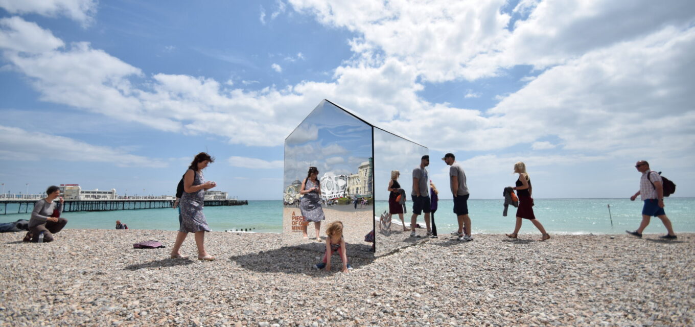 Beach with people walking around a beach hut made out of mirrors, reflecting the people and the surroundings