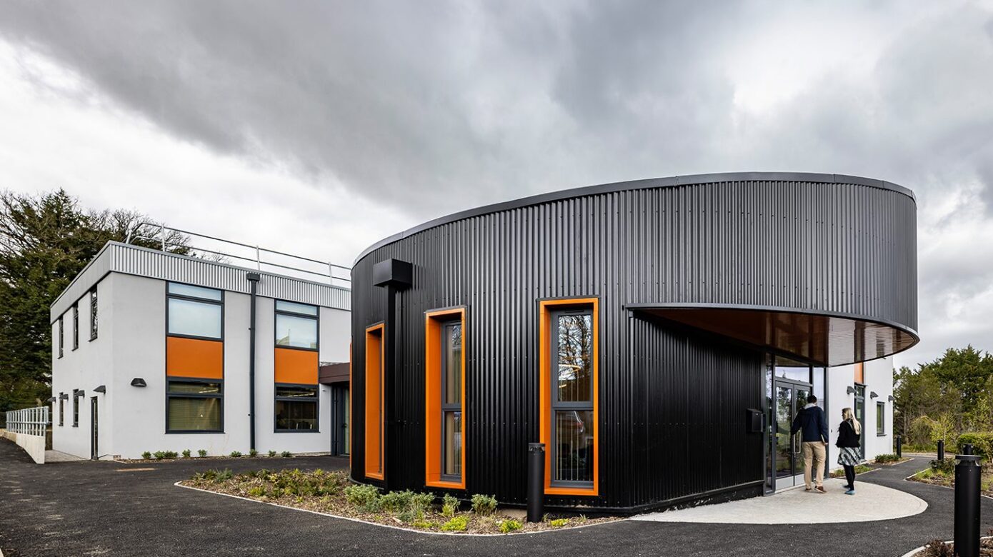 Large office entrance exterior clad in black metal, with orange window frames and a white extension to the back,