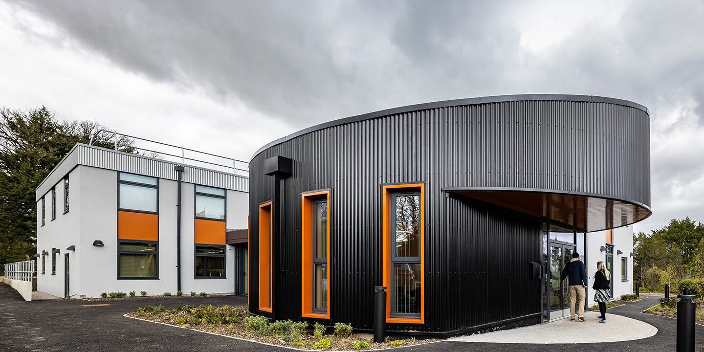 Large office entrance exterior clad in black metal, with orange window frames and a white extension to the back,