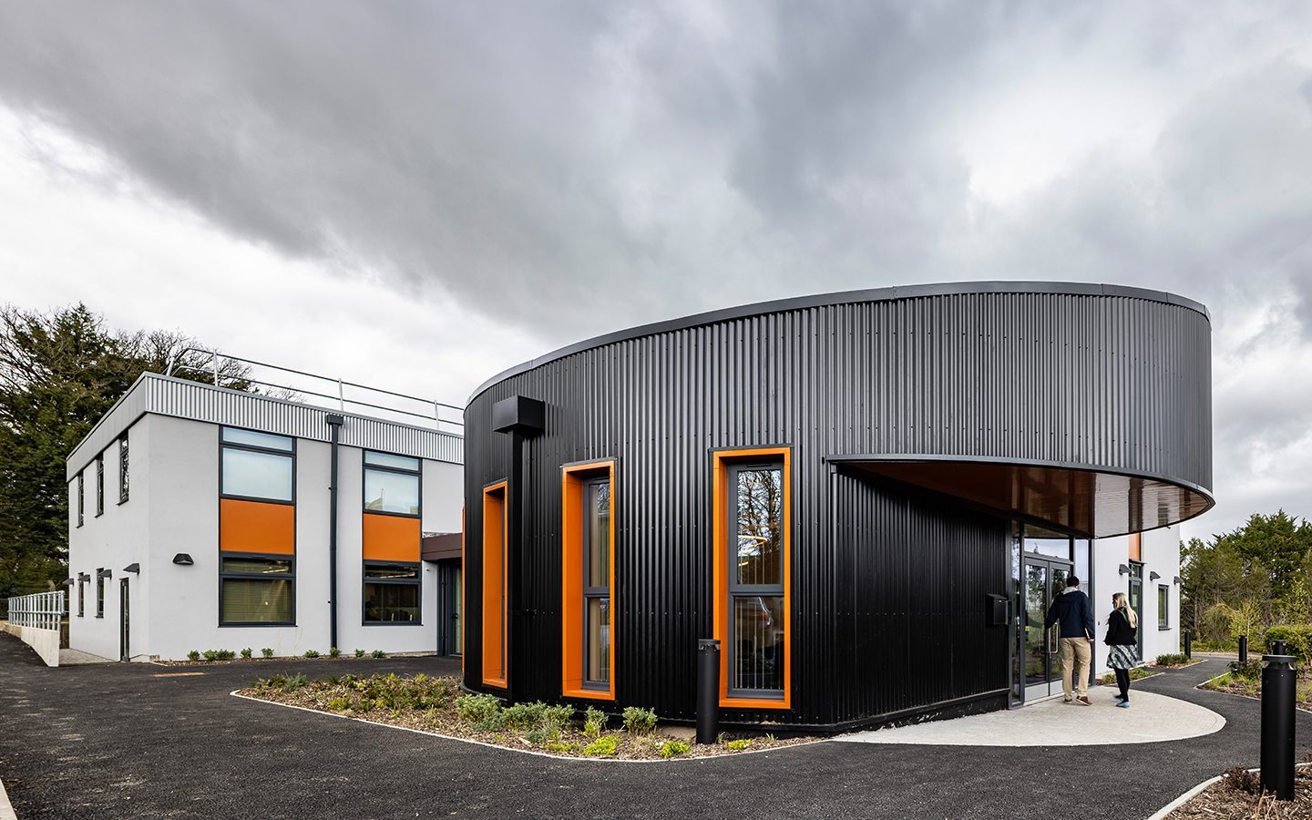 Large office entrance exterior clad in black metal, with orange window frames and a white extension to the back,