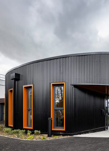 Large office entrance exterior clad in black metal, with orange window frames and a white extension to the back,