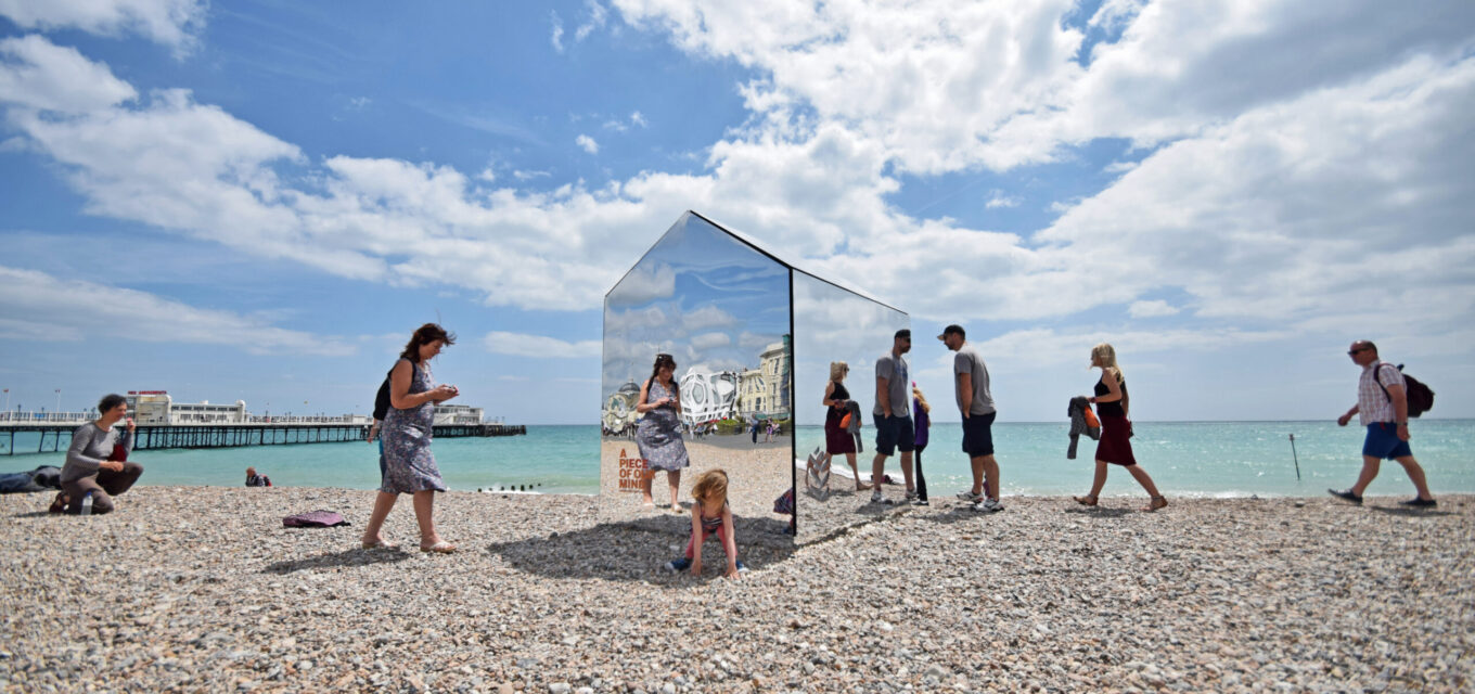 People surrounding a beach hut constructed out of mirrors, on a beach in Sussex