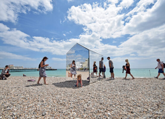 People surrounding a beach hut constructed out of mirrors, on a beach in Sussex